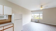 Located at 737 SW 17th Ave in Portland, Oregon, this studio unit offers a well-organized single-floor layout that efficiently combines the living, dining, and kitchen areas into one open space. Light-toned laminate flooring runs throughout, lending a clean and cohesive look to the interior. The combined living and dining area is brightened by a large window framing city and treetop views, complemented by a ceiling fan with integrated lighting. A wall-mounted HVAC unit provides climate control directly beneath the window. The kitchen is positioned along one wall in an efficient galley-style arrangement, featuring white cabinetry with ample upper and lower storage, a full-size refrigerator, an electric range, a built-in microwave, and a stainless steel sink. A dedicated closet with mirrored sliding doors provides practical storage within the main living space. The bathroom is outright functional, offering a single-sink vanity with a wide mirror, white cabinetry below, and a full tub-shower combination with a curtain rod. Neutral warm-beige walls throughout the unit create a calm, understated ambiance. The space is well-suited for those seeking a practical and efficiently designed home in a central Portland location.