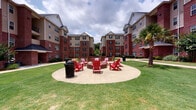 Located at 141 Hemlock Dr in Auburn, Alabama, this community clubhouse serves as a dynamic social hub set within a well-maintained multi-building residential complex. The exterior grounds are defined by classic red brick architecture, lush green lawns, and thoughtfully designed outdoor amenity spaces. A central courtyard features a circular fire pit lounge surrounded by bright red Adirondack chairs, perfect for gathering, while a sand volleyball court provides additional recreational options nearby.

Inside, the clubhouse boasts a bold and eclectic design characterized by a striking orange ceiling, exposed red brick accent walls, and rich polished concrete flooring. The main lounge area offers comfortable seating arranged around a brick fireplace, creating a warm and inviting atmosphere. Adjacent to the lounge, a full-service coffee and refreshment bar with dark cabinetry, pendant lighting, and bar stool seating serves as a central gathering point.

A dedicated dining area lines a wall of large windows fitted with bamboo shades and wooden blinds, allowing natural light to filter in while offering views of the surrounding outdoor spaces. Dark wood tables and woven leather chairs give the dining area a rustic, collegiate charm. The entryway features an exposed brick wall accented with coffee-themed décor, setting an energetic tone upon arrival. Overall, the space blends comfort, personality, and community-oriented design seamlessly.