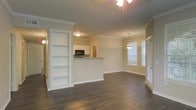 This single-floor space showcases an open-concept layout that seamlessly connects the living, dining, and kitchen areas. Dark hardwood flooring runs throughout, providing a rich contrast against the neutral, warm-toned walls and crisp white trim. Crown molding adds a refined architectural detail along the ceiling line, lending the space a polished, finished look.

A standout feature is the built-in bookshelf unit, recessed into the wall and painted white, offering both functional storage and a decorative focal point. Adjacent to it, a half-wall pass-through opens to the kitchen, which is equipped with a black microwave and white cabinetry, maintaining a clean, classic aesthetic.

The dining area is defined by a pendant light fixture that hangs elegantly above, while a ceiling fan with a light kit serves the main living area. Large windows dressed with white blinds allow generous natural light to fill the space, creating a bright and airy ambiance. A closeted door near the dining area adds practical storage. The overall design balances functionality with understated elegance throughout.