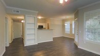 This single-floor space showcases an open-concept layout that seamlessly connects the living, dining, and kitchen areas. Dark hardwood flooring runs throughout, providing a rich contrast against the neutral, warm-toned walls and crisp white trim. Crown molding adds a refined architectural detail along the ceiling line, lending the space a polished, finished look.

A standout feature is the built-in bookshelf unit, recessed into the wall and painted white, offering both functional storage and a decorative focal point. Adjacent to it, a half-wall pass-through opens to the kitchen, which is equipped with a black microwave and white cabinetry, maintaining a clean, classic aesthetic.

The dining area is defined by a pendant light fixture that hangs elegantly above, while a ceiling fan with a light kit serves the main living area. Large windows dressed with white blinds allow generous natural light to fill the space, creating a bright and airy ambiance. A closeted door near the dining area adds practical storage. The overall design balances functionality with understated elegance throughout.