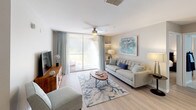 This single-floor residence is thoughtfully designed with a coastal-inspired palette of soft blues, warm neutrals, and crisp whites throughout. The open-concept living area features light hardwood-style flooring, a ceiling fan, and a large sliding glass door that floods the space with natural light and provides access to a private balcony. Adjacent to the living room, a dedicated dining area accommodates a four-seat table beneath a decorative sunburst mirror and an arched architectural niche.

The kitchen is outfitted with white shaker cabinetry, quartz countertops, a subway tile backsplash, stainless steel appliances, and a double-basin sink set into an island that opens toward the dining space. Tile flooring and ample storage complete the functional layout.

The primary bedroom features plush carpet, a ceiling fan, a walk-in closet, and an en-suite bathroom with a tub-shower combination and a white vanity. A second bedroom also offers carpet flooring, a ceiling fan, and generous natural light. The second full bathroom mirrors the clean, modern finishes found throughout. A dedicated laundry area with a pantry closet provides added convenience and storage.