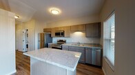 This single-floor unit offers a thoughtfully designed layout with contemporary finishes throughout. The kitchen serves as a central highlight, featuring warm-toned shaker-style cabinetry in a taupe-gray finish that extends to both upper and lower cabinets, providing generous storage. Light granite countertops with subtle veining complement the cabinetry and extend across a sizeable center island, ideal for meal preparation or casual dining. A full suite of stainless steel appliances — including a refrigerator, electric range, over-the-range microwave, and dishwasher — adds a polished, functional edge. Wood-look plank flooring runs throughout the main living areas, offering durability and a cohesive aesthetic. A wall-mounted mini-split unit provides efficient climate control in the open living area. A sliding glass door leads to a balcony, introducing natural light and outdoor access. The hallway connects the living spaces to additional rooms, with a dedicated electrical panel access point along the corridor. The bathroom features wood-look tile flooring, a vanity with storage, and a clean, neutral palette that maintains the unit's consistent modern character.