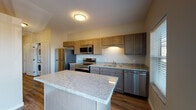 This single-floor unit offers a thoughtfully designed layout with contemporary finishes throughout. The kitchen serves as a central highlight, featuring warm-toned shaker-style cabinetry in a taupe-gray finish that extends to both upper and lower cabinets, providing generous storage. Light granite countertops with subtle veining complement the cabinetry and extend across a sizeable center island, ideal for meal preparation or casual dining. A full suite of stainless steel appliances — including a refrigerator, electric range, over-the-range microwave, and dishwasher — adds a polished, functional edge. Wood-look plank flooring runs throughout the main living areas, offering durability and a cohesive aesthetic. A wall-mounted mini-split unit provides efficient climate control in the open living area. A sliding glass door leads to a balcony, introducing natural light and outdoor access. The hallway connects the living spaces to additional rooms, with a dedicated electrical panel access point along the corridor. The bathroom features wood-look tile flooring, a vanity with storage, and a clean, neutral palette that maintains the unit's consistent modern character.