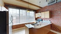 This single-floor residence on 17th Street combines industrial character with warm, inviting design elements throughout its layout. A long central hallway serves as the spine of the space, lined with smooth white walls contrasting against rich hardwood flooring that extends throughout. Exposed wooden ceiling beams and visible utility conduits running along the ceiling add an industrial, loft-like aesthetic, while natural light filters in from the far end, creating a bright and welcoming atmosphere.

The kitchen is a standout feature, showcasing full exposed brick walls that lend a rugged, urban charm to the space. Light wood cabinetry pairs well with granite countertops, and a peninsula with an integrated double sink provides both additional workspace and a casual gathering area. A stainless electric range and over-range ventilation hood offer practical cooking functionality. Large windows fitted with horizontal blinds allow abundant natural light into the kitchen while framing views of the surrounding urban landscape. Pendant lighting above the peninsula adds a polished finishing touch. The combination of brick, wood beams, and hardwood floors gives the overall space a cohesive, character-rich aesthetic.