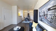 This single-floor space showcases a contemporary open-concept design that connects the living and dining areas in a cohesive, functional layout. Dark hardwood flooring runs throughout, providing a rich contrast to the crisp white walls and lending a polished, modern feel to the interior.

The living area is anchored by a leather sofa accented with yellow and grey throw pillows, complemented by a sleek black coffee table. A large black-and-white photographic art piece adorns the wall, adding a sophisticated, urban aesthetic to the space. A tall dark bookcase provides additional storage and visual depth.

The dining area, positioned toward the rear of the open layout, features a dark wood dining table with matching chairs set atop a light area rug. A chalkboard panel serves as a decorative backdrop, adding a casual, creative touch. Track lighting overhead ensures the dining space is well-lit and inviting.

A pass-through opening hints at an adjacent kitchen, which features warm yellow accents visible from the main living area. The overall ambiance is modern and well-appointed, with a neutral color palette punctuated by bold accent tones throughout.