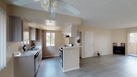 This single-story unit presents a practical and straightforward layout designed for comfortable everyday living. The open-concept common area connects the kitchen and living room seamlessly, with light gray wood-look flooring running throughout these shared spaces, contributing to a clean, cohesive aesthetic. Warm neutral tones on the walls complement the light flooring and create a bright, welcoming atmosphere throughout.

The kitchen is efficiently arranged in a galley-style configuration, featuring white cabinetry, a double-basin sink, a gas range, and an under-cabinet range hood. A raised peninsula provides a natural divider between the kitchen and living area. A window above the counter allows natural light to fill the space. The kitchen ceiling includes a distinct tray detail with a drop panel, adding a subtle architectural element.

The living room is generously proportioned, with a ceiling fan overhead and large sliding glass doors fitted with vertical blinds that open to an outdoor patio, allowing ample natural light. A wall-mounted heating and cooling unit provides climate control.

The bedrooms feature plush carpeting, textured ceilings, and windows or sliding door access to outdoor views. The bathroom includes standard fixtures in a compact layout. Overall, the unit offers a functional and well-maintained living environment.