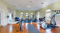 This fitness center offers a generously sized, single-floor workout space thoughtfully designed to accommodate a wide range of exercise routines. The room is immediately distinguished by its eye-catching green and white patterned wallpaper, which lends a unique and inviting character to the space. Elegant arched transom windows line the walls, allowing ample natural light to flood the interior and creating an open, energizing atmosphere.

The flooring combines warm hardwood with rubberized exercise mats, delineating dedicated zones for cardio, strength training, and stretching. The cardio section is well-appointed with multiple treadmills, elliptical machines, a rowing machine, and spin bikes. The strength training area features a multi-station cable machine, a dumbbell rack with a full range of weights, and various benches and attachments for a complete workout.

A convenient amenity area includes a beverage vending machine, a small kitchenette with cabinetry and a sink, ideal for post-workout refreshment. Additional features include a wall-mounted television, stacked exercise mats for group fitness use, and recessed ceiling lighting complemented by flush-mount fixtures. The space is accessible via multiple entry points, including an exterior door that welcomes in natural views, reinforcing the bright and welcoming ambiance throughout.