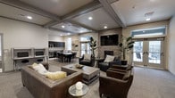 This well-appointed single-floor space is designed for comfort and socializing, blending modern aesthetics with warm, inviting finishes throughout. The main lounge area serves as the centerpiece, anchored by a striking stacked stone fireplace wall with a mounted flat-screen television above. Plush leather sofas and accent chairs are arranged around a rustic wooden coffee table, creating an ideal setting for casual gatherings or relaxed conversation. Tall indoor plants add a refreshing natural element to the space.

Overhead, coffered ceilings with recessed lighting lend an architectural depth and a sense of sophistication to the room, while the neutral tones of the walls and flooring provide a cohesive, modern backdrop. Natural light filters in through multiple windows, brightening the interior and offering views to the outdoors.

Flowing seamlessly from the lounge is a dedicated dining area, furnished with a large table and chairs to accommodate group meals or meetings. Adjacent to the dining space is a well-equipped kitchenette featuring dark cabinetry and modern appliances, making it convenient for hosting and entertaining. The overall ambiance is polished yet welcoming, making this a versatile and functional communal space.