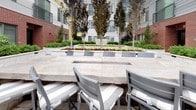 Located at 180R Cambridgepark Dr in Cambridge, Massachusetts, this thoughtfully designed outdoor courtyard offers a welcoming communal retreat. The space features large-format stone tile flooring in a neutral palette, lending a clean and polished aesthetic underfoot. A gracefully curved stone retaining wall defines the seating area, doubling as a planter that supports lush, layered landscaping filled with ornamental shrubs, ground cover, and small trees. The greenery adds year-round texture and a sense of natural enclosure within the urban setting.

Cushioned outdoor chairs and generous stone-topped bar-style tables provide ample seating for gathering and dining al fresco. A dedicated outdoor kitchen station anchors one end of the courtyard, equipped with a stainless steel gas grill set into a refined stone surround, making it well-suited for outdoor entertaining. Upright ornamental trees rise throughout the central planting beds, adding vertical interest and a sense of privacy from the surrounding multi-story building facades, which feature a complementary mix of brick, panel siding, and large windows. The overall ambiance is refined yet approachable, blending durable materials with abundant greenery to create an inviting outdoor environment ideal for relaxation and socializing.