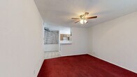 Located at 1402 Fannidella Dr in Carrollton, Texas, this single-story 2-bedroom, 2-bathroom home offers a practical and well-organized layout. The living room is carpeted in a rich, deep red and features a ceiling fan with light fixture, white-painted walls, and a window with blinds that allows natural light to filter in. It connects openly to the kitchen via a pass-through counter, supporting a convenient flow between spaces. The galley-style kitchen is equipped with warm wood-toned cabinetry, a double-basin sink, a gas range, a refrigerator, and tile flooring, with a window above the sink providing additional natural light. Both bedrooms continue the deep red carpet theme, each fitted with ceiling fans and windows with blinds. The primary bedroom offers a notably generous footprint, while the second bedroom includes a sliding-door closet with built-in shelving. Both bathrooms feature tub-shower combinations with light-toned tile surrounds, stone-pattern tile flooring, and standard white fixtures. A hallway connects the main living areas to the bedrooms and bathrooms, keeping the layout straightforward and functional throughout.