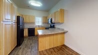 This single-story residence offers a well-organized layout that connects living, dining, and kitchen spaces in an open, functional arrangement. The entry transitions from tile flooring into a carpeted living area, where vaulted ceilings add a sense of height and openness. A door with sidelight windows provides access to the outdoors, bringing in natural light and contributing to the airy feel of the space.

The kitchen is a standout feature, equipped with light wood cabinetry, granite-patterned countertops, and a full suite of black appliances including a refrigerator, range, dishwasher, and over-the-range microwave. A peninsula with a curved granite counter extends into the dining area, offering additional prep space and casual seating. Wood-look flooring runs throughout the kitchen and dining zone, while a window above the sink provides natural light. An elegant chandelier overhead enhances the dining atmosphere.

Bedrooms are carpeted and include sliding or hinged closet doors with built-in hanging rods. Both bathrooms are finished consistently with granite-top vanities, light wood cabinetry, tile flooring, and tub-shower combinations, providing a cohesive aesthetic throughout the home.