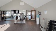 Fitness Center 2 is a thoughtfully designed exercise facility that combines an open, airy atmosphere with a comprehensive selection of workout equipment. The space is defined by its striking vaulted ceiling, which rises dramatically to create a sense of openness and volume throughout the room. Large windows line the walls, flooding the interior with natural light and providing views of the surrounding outdoor landscape, even in winter conditions.

The flooring consists of durable dark rubber gym flooring, providing both safety and comfort for a wide range of exercises. The cardio section is well-appointed with multiple treadmills, an elliptical trainer, and a stationary bike, all positioned to take advantage of the natural light from the surrounding windows. A dedicated strength training area includes a cable machine, a leg press machine, and a rack of dumbbells, catering to a variety of resistance training needs.

A large wall-mounted mirror along one side of the room enhances the sense of space and supports proper form during workouts, and a flat-screen television is mounted nearby for entertainment. Additional amenities include a water cooler station for hydration and a dumbbell rack with neatly organized free weights. Recessed lighting complements the natural light, ensuring the space remains well-lit at all times. The facility is accessible via a glass-paneled entry door and is clearly marked with safety signage.
