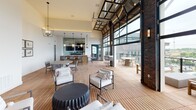 This sophisticated rooftop lounge blends industrial character with modern comfort across a single, open-concept floor. The space is anchored by a dramatic exposed brick accent wall that rises to soaring heights, housing a built-in gas fireplace topped by a mounted flat-screen television and a rustic wood mantel. Warm teak deck flooring flows throughout the interior and extends seamlessly onto the open-air terrace, reinforcing a cohesive indoor-outdoor aesthetic.

Floor-to-ceiling glass garage doors with bold black steel framing serve as the defining architectural feature, folding upward to dissolve the boundary between the enclosed lounge and the expansive outdoor terrace beyond. The terrace is bathed in natural light and offers sweeping panoramic views of the surrounding cityscape and a nearby ballpark, making it an exceptional vantage point for gathering and entertaining.

Inside, multiple seating arrangements invite relaxation, including woven lounge chairs, cushioned benches, and low concrete accent tables arranged around the fireplace. A built-in bar and kitchen area with pendant lighting and a wall-mounted TV provides a hub for hosting. A pool table and additional mounted screen offer recreational amenities. Wall sconces and chandelier-style pendant fixtures provide warm, layered lighting throughout, completing this dynamic and well-appointed communal space.