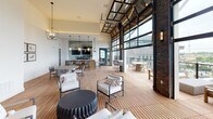 This sophisticated rooftop lounge blends industrial character with modern comfort across a single, open-concept floor. The space is anchored by a dramatic exposed brick accent wall that rises to soaring heights, housing a built-in gas fireplace topped by a mounted flat-screen television and a rustic wood mantel. Warm teak deck flooring flows throughout the interior and extends seamlessly onto the open-air terrace, reinforcing a cohesive indoor-outdoor aesthetic.

Floor-to-ceiling glass garage doors with bold black steel framing serve as the defining architectural feature, folding upward to dissolve the boundary between the enclosed lounge and the expansive outdoor terrace beyond. The terrace is bathed in natural light and offers sweeping panoramic views of the surrounding cityscape and a nearby ballpark, making it an exceptional vantage point for gathering and entertaining.

Inside, multiple seating arrangements invite relaxation, including woven lounge chairs, cushioned benches, and low concrete accent tables arranged around the fireplace. A built-in bar and kitchen area with pendant lighting and a wall-mounted TV provides a hub for hosting. A pool table and additional mounted screen offer recreational amenities. Wall sconces and chandelier-style pendant fixtures provide warm, layered lighting throughout, completing this dynamic and well-appointed communal space.