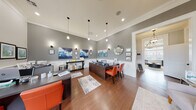 This single-floor professional office space is thoughtfully designed to balance functionality with an inviting, upscale aesthetic. The main reception and workspace area serves as the heart of the office, featuring rich hardwood flooring, crisp white wainscoting, and walls finished in a sophisticated gray tone. Multiple dark-stained workstations are paired with vibrant orange leather chairs, creating a bold yet professional contrast that energizes the space. A series of globe pendant lights hang at varying heights from the elevated ceiling, supplemented by recessed lighting and wall-mounted sconces that ensure warm, even illumination throughout. Decorative artwork, including blue abstract canvas pieces and a cityscape print, adds visual interest to the walls alongside a round mirror accent. A welcoming lounge area near the entrance features tufted upholstered chairs and a small accent table, offering a comfortable space for guests. Natural light filters through windows fitted with clean white blinds. A private interior office provides a more focused work environment, complete with an L-shaped desk, filing storage, and additional natural light. Connecting hallways with elegant pendant lighting lead between the distinct functional zones, giving the overall layout a refined sense of flow and professionalism.