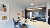 This single-floor 1-bedroom, 1-bathroom residence showcases a thoughtfully designed open-concept layout that seamlessly integrates the kitchen and dining areas. The kitchen is outfitted with stainless steel appliances, including a full-size refrigerator and a built-in microwave, complemented by dark espresso cabinetry that extends to the ceiling for ample storage. A textured tile backsplash adds depth and contrast against the rich cabinetry tones. The light-colored countertop extends outward to form a breakfast bar, providing casual seating for two with modern high-back chairs — a practical and stylish dining solution. Under-cabinet LED strip lighting and a suspended pendant fixture above the bar illuminate the workspace effectively. The overall color palette balances warm and cool tones, with crisp white walls contrasting against the dark cabinetry. Decorative touches, including a framed artwork piece and fresh floral arrangements, contribute to the inviting ambiance. The clean lines and contemporary finishes throughout create a cohesive, modern aesthetic well-suited for efficient urban living.