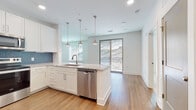 Located at 1040 Enclave Piney Lane in Asheville, NC, this thoughtfully designed single-floor residence combines modern finishes with a clean, contemporary aesthetic throughout. The open-concept kitchen is a standout feature, showcasing crisp white shaker cabinetry that extends to the ceiling, granite countertops, and a striking blue subway tile backsplash. A large central island with an undermount sink and pendant lighting serves as the heart of the space, complemented by a full suite of stainless steel appliances including a French door refrigerator, electric range, microwave, and dishwasher. Warm hardwood-style flooring flows seamlessly through the kitchen and into the living area, which is anchored by a ceiling fan and a set of sliding glass doors opening onto a private balcony with picturesque wooded hillside views. The bedrooms offer plush carpeting and generous natural light from multiple windows. The bathrooms are finished with the same attention to detail, featuring white vanities with granite countertops, large mirrors, and hardwood-style flooring. One bathroom includes a glass-enclosed shower and direct access to a walk-in closet. Recessed lighting throughout, neutral tones, and a cohesive design palette give the entire space a fresh, move-in-ready feel.