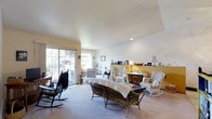 This single-floor space offers a warm and inviting atmosphere centered around a generously sized living room that serves as the heart of the home. The living room is bathed in natural light streaming through large sliding glass doors that open onto an outdoor balcony, creating a seamless connection between the interior and exterior. The smooth white ceiling features subtle recessed lighting, adding a clean, bright feel to the room. Neutral wall-to-wall carpeting runs throughout, lending a soft and comfortable underfoot experience. The space is characterized by its classic, cottage-inspired charm, with an eclectic mix of wicker furniture, rocking chairs, and draped seating arrangements that contribute to a relaxed, lived-in ambiance. A warm golden-yellow accent wall in the background adds a touch of color and depth, complementing the otherwise neutral palette of white and cream tones. Decorative shelving and personal accents, including framed photos, candles, and small ornaments, are thoughtfully arranged throughout, giving the space a personal and homey character. The open layout allows for comfortable movement and flexible furniture arrangement, making it a functional and welcoming environment suited for both relaxing and entertaining.