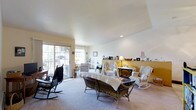 This single-floor space offers a warm and inviting atmosphere centered around a generously sized living room that serves as the heart of the home. The living room is bathed in natural light streaming through large sliding glass doors that open onto an outdoor balcony, creating a seamless connection between the interior and exterior. The smooth white ceiling features subtle recessed lighting, adding a clean, bright feel to the room. Neutral wall-to-wall carpeting runs throughout, lending a soft and comfortable underfoot experience. The space is characterized by its classic, cottage-inspired charm, with an eclectic mix of wicker furniture, rocking chairs, and draped seating arrangements that contribute to a relaxed, lived-in ambiance. A warm golden-yellow accent wall in the background adds a touch of color and depth, complementing the otherwise neutral palette of white and cream tones. Decorative shelving and personal accents, including framed photos, candles, and small ornaments, are thoughtfully arranged throughout, giving the space a personal and homey character. The open layout allows for comfortable movement and flexible furniture arrangement, making it a functional and welcoming environment suited for both relaxing and entertaining.