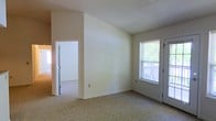 This single-floor residential space offers a clean, functional layout with a straightforward design. The main living area is finished with wall-to-wall carpeting in a neutral beige tone, complementing the light cream-colored walls that create a bright and understated atmosphere throughout. A textured ceiling adds a subtle architectural detail to the space.

Natural light enters through a window fitted with horizontal blinds, offering a view of an outdoor deck or porch with a railing visible beyond the glass — a pleasant feature that connects the interior to the outdoors. Adjacent to the window, a doorway provides direct access to this exterior area.

An interior doorway leads to a well-lit adjoining room, suggesting a connected layout that allows for easy movement between spaces. The walls feature clean white trim and baseboards, providing a crisp contrast to the warmer wall tones. A standard electrical outlet is visible along the lower wall, reflecting the practical, ready-to-use nature of the space.

Overall, the space presents a modest and functional residential environment with neutral finishes, natural light, and a layout designed for comfortable everyday living. The uncluttered interior offers a blank canvas that can be easily personalized to suit a variety of needs and styles.