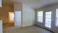 This single-floor residential space offers a clean, functional layout with a straightforward design. The main living area is finished with wall-to-wall carpeting in a neutral beige tone, complementing the light cream-colored walls that create a bright and understated atmosphere throughout. A textured ceiling adds a subtle architectural detail to the space.

Natural light enters through a window fitted with horizontal blinds, offering a view of an outdoor deck or porch with a railing visible beyond the glass — a pleasant feature that connects the interior to the outdoors. Adjacent to the window, a doorway provides direct access to this exterior area.

An interior doorway leads to a well-lit adjoining room, suggesting a connected layout that allows for easy movement between spaces. The walls feature clean white trim and baseboards, providing a crisp contrast to the warmer wall tones. A standard electrical outlet is visible along the lower wall, reflecting the practical, ready-to-use nature of the space.

Overall, the space presents a modest and functional residential environment with neutral finishes, natural light, and a layout designed for comfortable everyday living. The uncluttered interior offers a blank canvas that can be easily personalized to suit a variety of needs and styles.