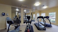 This fitness center is housed within a single-level space designed to accommodate a comprehensive workout experience. The room is finished with neutral-toned carpet flooring and warm yellow walls, creating an inviting atmosphere brightened by multiple windows that allow natural light to fill the space. Flush-mount ceiling light fixtures and ceiling fans provide additional illumination and air circulation throughout.

The cardio area is equipped with treadmills and elliptical trainers, positioned near the windowed wall to allow outdoor views during workouts. A multi-station cable machine and a pull-up/dip tower station serve as the centerpieces of the strength training section, complemented by a dedicated free weight area featuring a full dumbbell rack and a flat bench. Protective floor mats are placed beneath the equipment for safety and stability.

Full-length mirrors line one wall, visually expanding the space and allowing users to monitor their form. A wall-mounted flat-screen television adds convenience for entertainment. Along one side of the room, built-in wood cabinetry with a countertop provides a functional surface, likely used for storage or a refreshment station. A dedicated door provides direct exterior access, enhancing convenience for users entering and exiting the facility.