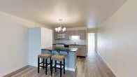 This single-floor residence offers a well-designed layout that blends functionality with contemporary style. The open-concept kitchen serves as a central feature, showcasing gray cabinetry paired with a classic white subway tile backsplash. White appliances, including a range and over-the-range microwave, are seamlessly integrated into the cabinetry. A peninsula with a dark countertop extends into the living area, providing seating for two with upholstered bar stools, making it ideal for casual dining or entertaining. A chandelier with candelabra-style lights hangs above, adding a touch of elegance to the space. The kitchen also includes a stainless steel sink positioned beneath upper cabinetry, with ample counter space on either side. Warm-toned luxury vinyl plank flooring runs throughout, contributing to a cohesive and clean aesthetic. The open sightlines extend toward a hallway leading to additional rooms, including the bedroom and bathroom. Light-toned walls throughout keep the space feeling bright and airy. Overall, the residence presents a practical and visually appealing interior well-suited for comfortable everyday living.