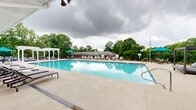 Located at 201 Water Hill Rd in Madison, Alabama, this six-floor community offers a well-appointed outdoor amenity area designed for relaxation and recreation. The centerpiece of the outdoor space is a generously sized swimming pool with vivid blue water, framed by a wide concrete deck lined with rows of sling-style lounge chairs ideal for sunbathing. A classic white pergola anchors one corner of the pool area, providing a shaded gathering spot, while teal-accented umbrellas add pops of color throughout.

Backing the pool is a charming brick clubhouse building featuring a steeply pitched gray roof, skylights, and French doors that open directly onto the pool deck — creating a seamless indoor-outdoor connection. The clubhouse provides convenient access to restrooms and additional amenities.

On the opposite side of the pool, a spacious patio area offers casual outdoor seating with high-top tables and chairs, as well as a ping-pong table for recreation. The entire pool and patio area is enclosed by a black iron fence, ensuring privacy and security while maintaining open views of the surrounding mature trees and lush greenery. The natural landscaping creates a tranquil, park-like atmosphere that enhances the overall outdoor experience of this Madison community.