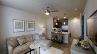 This single-floor residence offers a thoughtfully designed open-concept layout that seamlessly connects the living area and kitchen, creating a spacious and functional living environment. The living area is warm and inviting, featuring wood-look flooring, neutral toned walls, and a ceiling fan with integrated lighting that enhances both comfort and style. A comfortable sectional sofa, decorative accent pillows, and carefully selected wall art contribute to the cozy yet refined atmosphere. A plush area rug anchors the seating area, adding texture and warmth to the space. The kitchen is a standout feature, showcasing dark cabinetry, stainless steel appliances, and a light-toned stone countertop that extends into a peninsula breakfast bar. Woven rattan bar stools provide casual seating at the bar, making it an ideal spot for dining or entertaining. Elegant pendant lights hang above the peninsula, complementing the recessed lighting throughout. The kitchen and living areas flow naturally into one another, making the space feel open and well-connected. The overall design balances modern functionality with comfortable, residential warmth, making it a practical and visually appealing place to call home.