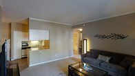 This single-floor residence offers a thoughtfully designed layout that blends comfort and functionality. The living area serves as the heart of the space, featuring plush wall-to-wall carpeting, warm neutral-toned walls, and crisp crown molding that adds a polished architectural touch to the room. Ambient lighting enhances the cozy atmosphere, complementing the understated décor. A sleek glass-top coffee table and a contemporary sofa anchor the living space, creating an inviting area for relaxation.

A pass-through kitchen window connects the living area to the kitchen, providing an open, communicative feel between the two spaces. The kitchen is equipped with a sink and cabinetry finished in warm wood tones, maximizing efficiency within a compact footprint.

From the living area, a hallway leads to additional rooms, including what appears to be a bathroom with a bathtub and built-in shelving visible through the open doorway. A tall, illuminated floor lamp positioned in the hallway casts a warm, welcoming glow throughout the passage.

The overall ambiance of this residence is calm and neutral, with consistent finishes and thoughtful lighting creating a cohesive, comfortable living environment throughout the single floor.