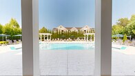 Located at 30 Gates Mill St in Huntsville, Alabama, this two-story community amenity space is designed to offer a resort-like outdoor experience within a well-maintained residential setting. At the heart of the property is a generously sized swimming pool with crystal-clear water, in-pool lounge chairs, and stainless steel entry rails. The pool deck is finished in light stone pavers and outfitted with a variety of lounge chairs and umbrella-shaded seating, providing ample space for relaxation under the Alabama sun. Two white pergolas flank the pool area, adding architectural interest while creating shaded gathering spots along the perimeter. Adjacent to the pool, a dedicated outdoor lounge and dining area features multiple table-and-chair sets beneath large blue umbrellas, surrounded by lush, manicured landscaping including tall privacy hedges, neatly trimmed shrubs, and mature trees. A paved walking path winds through the greenery, connecting the various outdoor zones. The two-story clubhouse anchors the space with classic Southern architecture, featuring a red brick façade, white columned portico, dormer windows, and a pitched shingle roof. Tall, mature trees frame the entire property, providing natural shade and a sense of seclusion throughout the amenity grounds.