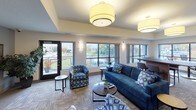 This single-floor community space is thoughtfully designed to accommodate a variety of activities across several distinct functional zones. The lounge area serves as the centerpiece, furnished with a plush navy blue sofa and coordinating geometric-patterned accent chairs arranged around a round coffee table. Overhead, large drum pendant light fixtures provide warm, even illumination throughout. A dedicated entertainment zone features a wall-mounted flat-screen television flanked by framed artwork and lush indoor plants, creating an inviting atmosphere for gathering and relaxation. Adjacent to the lounge, a study or work area offers a large, dark-surfaced table with bar-height seating, ideal for collaborative work or individual tasks. This zone benefits from generous natural light streaming through a wide bank of windows that frame outdoor views of surrounding greenery and the surrounding neighborhood. The kitchen is fully equipped with rich dark-stained cabinetry, stainless steel appliances including a refrigerator, range, and microwave, and a granite-topped breakfast bar with bar stools. Neutral-toned carpeting and tray ceilings with recessed lighting add architectural interest throughout, while wall sconces contribute to the warm, welcoming ambiance of the overall space.