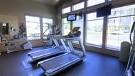 This fitness center offers a bright and functional workout environment designed to support a range of cardiovascular exercise needs. Large windows line the walls on multiple sides, flooding the space with natural light and providing open views of the surrounding outdoor landscape, creating an airy and motivating atmosphere. The room features rubber flooring suited for high-traffic gym use, providing both durability and comfort underfoot. A row of modern treadmills serves as the centerpiece of the space, each equipped with individual digital display consoles for tracking workout metrics. Behind them, elliptical-style cross-trainers and additional strength or cable equipment add variety to the available workout options. Wall-mounted flat-screen televisions are positioned at multiple points for easy viewing from the cardio equipment, enhancing the exercise experience. A wall-mounted fan provides supplemental ventilation. The high placement of windows near the roofline contributes to an elevated, open feel within the room. Overall, the space is clean, well-lit, and thoughtfully arranged to maximize both equipment accessibility and comfort for users.