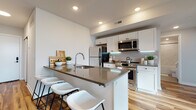 This contemporary single-floor residence showcases a thoughtfully designed open-concept layout that seamlessly connects the main living areas. The kitchen is a standout feature, outfitted with crisp white cabinetry, a herringbone-tiled backsplash, stainless steel appliances, and a spacious island with a dark quartz countertop and bar seating — ideal for both cooking and casual dining. Warm-toned hardwood flooring runs throughout the living and dining areas, enhancing the bright, airy ambiance created by recessed lighting and generous natural light. The living room is anchored by dramatic floor-to-ceiling windows framed in matte black, offering expansive urban views and flooding the space with daylight. The bedrooms continue the theme of abundant natural light, each featuring large black-framed windows with city views. Both bathrooms are finished with marble-style tile surrounds and matte black fixtures — one featuring a glass-enclosed walk-in shower, the other a full soaking tub. Clean white walls, modern hardware, and a cohesive neutral palette throughout give this residence a polished, contemporary character that is both stylish and functional.