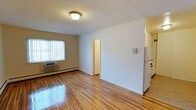 Located at 780 Rockaway Avenue in the Maple Crest neighborhood of Valley Stream, NY 11581, this single-floor residence offers a clean and functional layout designed for comfortable everyday living. The main living area is highlighted by gleaming hardwood floors that reflect natural light streaming in through a large window fitted with vertical blinds, creating a bright and welcoming atmosphere. The room is well-proportioned, providing ample open space for furnishing and personalization. A wall-mounted thermostat and a baseboard heating unit with an integrated window air conditioner ensure year-round climate control. The living area flows naturally toward a doorway that leads to additional spaces within the unit. Adjacent to the living room, the kitchen is partially visible and includes a full-sized refrigerator, with cabinetry providing practical storage. The neutral color palette throughout — warm beige walls and white trim — lends the space a clean, move-in-ready appearance. Flush-mount ceiling lighting provides consistent illumination throughout. Overall, this unit offers a straightforward, well-maintained living environment in a convenient Valley Stream location.