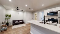 This well-designed single-floor space offers a seamless blend of modern aesthetics and practical functionality. The open-concept layout connects the kitchen and living area, creating a cohesive and comfortable environment. The kitchen is a standout feature, equipped with stainless steel appliances including an electric range, over-the-range microwave, and refrigerator. Crisp white shaker-style upper cabinets are paired with gray lower cabinets, accented by matte black hardware. A peninsula with a bright white quartz countertop and an undermount double sink fitted with a sleek black faucet extends into the living space, providing additional prep and seating space. A mosaic tile backsplash adds a refined touch to the cooking area. The living space features wood-look flooring that runs throughout, neutral light gray walls, and recessed lighting complemented by a modern black ceiling fan, lending the room a bright and airy feel. The bathroom continues the clean, contemporary theme with full white subway tile surrounding a soaking tub and shower combination, paired with matte black fixtures and fittings. Recessed lighting illuminates the space consistently across all rooms, and interior doors feature matching black hardware, tying the design together cohesively.