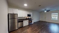 Located at 19 Clinton Ave in Bay Shore, NY, this single-floor unit at Graymore Flats offers a thoughtfully designed layout with a consistent, modern aesthetic throughout. Dark hardwood-style flooring runs through the main living areas, providing a warm contrast against the crisp white walls and creating a polished, cohesive look.

The open living area is spacious and well-lit, featuring a ceiling fan with integrated lighting and recessed fixtures that illuminate the room evenly. The space flows naturally toward the kitchen, which is outfitted with white shaker-style cabinetry, dark countertops, and a full suite of stainless steel appliances including a gas range, over-the-range microwave, dishwasher, and refrigerator. A window in the kitchen area brings in natural light.

The bedroom features soft carpeting and access to a generously sized walk-in closet with built-in shelving and rail systems, offering ample storage. The bathroom is cleanly finished with light tile flooring, a white vanity with a dark countertop, an oval mirror, and a full tub-shower combination with subway tile surround. Overall, the unit presents a clean, move-in-ready interior with functional, modern finishes throughout.
