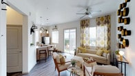 This single-story space offers an open-concept layout that seamlessly connects the living area and kitchen, creating a cohesive and functional environment. The living area is well-lit, benefiting from abundant natural light streaming through sliding glass doors that provide direct access to an outdoor patio, as well as additional windows dressed with decorative leaf-patterned curtains in warm yellow and brown tones. A ceiling fan overhead adds both practicality and a casual aesthetic to the space.

The kitchen is compact yet well-appointed, featuring wood-tone cabinetry, a stainless steel refrigerator, and pendant lighting suspended above a breakfast bar with barstool seating. Open shelving adds a modern touch while keeping the space feeling airy.

The living area is styled with a neutral-toned sofa, accent armchairs, and a glass-top coffee table, all arranged to encourage conversation and comfort. Decorative touches, including a mounted antler accent and potted greenery, contribute warmth and character. Durable wood-look flooring runs throughout, tying the spaces together cohesively while offering easy maintenance.