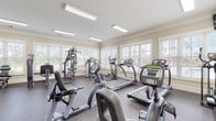 Located at 101 Tomshire Dr in Johns Island, South Carolina, this well-equipped fitness center offers a comprehensive workout environment designed to accommodate a wide range of exercise routines. The single-floor facility features a spacious, open layout with rubber-floored workout areas that provide both comfort and safety during exercise.

The cardio section is outfitted with multiple treadmills, elliptical trainers, and stationary bikes, all positioned along the perimeter walls where large, white-framed windows with blinds allow an abundance of natural light to fill the space and provide views of the surrounding outdoors. Overhead, flush-mounted fluorescent lighting ensures consistent illumination throughout.

The strength training area includes a multi-function cable machine, a weight bench with adjustable settings, a full rack of dumbbells in graduated sizes, and kettlebells — catering to both beginners and experienced athletes. Crown molding and clean white walls lend a polished, residential feel to the facility. A wall-mounted television is available for entertainment during workouts.

Elegant white French doors with glass panes connect to an adjacent room, adding an architectural charm uncommon in typical fitness spaces. Safety equipment, including a fire extinguisher, is also present. The overall ambiance is bright, clean, and functional, making this fitness center a well-rounded amenity.