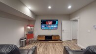 Located at 4890 Edwards Farm Rd in Hilliard, Ohio, this single-floor space is designed as a dedicated home theater and entertainment room. The room is finished with light wood-look luxury vinyl plank flooring and neutral gray walls, creating a clean, modern backdrop for the entertainment setup.

The focal point of the space is a large wall-mounted flat-screen television positioned at optimal viewing height, complemented by a stylish media console below featuring a warm wood-tone cabinet with black metal legs for media component storage. Recessed lighting in the ceiling provides well-distributed, adjustable illumination throughout the room, allowing for the ideal viewing atmosphere.

Plush, dark leather reclining theater-style seats with built-in cup holders are arranged to face the screen, delivering a true cinema-style experience. Adding to the authentic movie-night ambiance is a classic red commercial-style popcorn machine on a wheeled cart, positioned in the corner of the room as both a functional and decorative accent.

The space is accessed through a white paneled door with a modern keypad entry lock, providing both convenience and security. Overall, this thoughtfully designed entertainment space offers a comfortable, immersive home theater experience suited for movie nights and gatherings alike.
