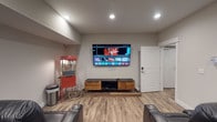 Located at 4890 Edwards Farm Rd in Hilliard, Ohio, this single-floor space is designed as a dedicated home theater and entertainment room. The room is finished with light wood-look luxury vinyl plank flooring and neutral gray walls, creating a clean, modern backdrop for the entertainment setup.

The focal point of the space is a large wall-mounted flat-screen television positioned at optimal viewing height, complemented by a stylish media console below featuring a warm wood-tone cabinet with black metal legs for media component storage. Recessed lighting in the ceiling provides well-distributed, adjustable illumination throughout the room, allowing for the ideal viewing atmosphere.

Plush, dark leather reclining theater-style seats with built-in cup holders are arranged to face the screen, delivering a true cinema-style experience. Adding to the authentic movie-night ambiance is a classic red commercial-style popcorn machine on a wheeled cart, positioned in the corner of the room as both a functional and decorative accent.

The space is accessed through a white paneled door with a modern keypad entry lock, providing both convenience and security. Overall, this thoughtfully designed entertainment space offers a comfortable, immersive home theater experience suited for movie nights and gatherings alike.