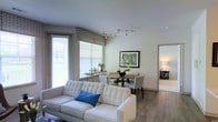 This single-floor space showcases a thoughtfully designed open-concept layout that seamlessly integrates the living and dining areas into one cohesive, functional space. The living area is anchored by a tufted white sofa accented with a vibrant blue decorative pillow, positioned to take advantage of the natural light streaming in through large windows dressed with patterned valances and horizontal blinds. The windows offer views of the surrounding outdoor greenery, adding a refreshing connection to the exterior environment.

Overhead, a sleek track lighting fixture provides adjustable, targeted illumination, complementing the neutral-toned walls and light hardwood-style flooring that extend throughout the space. The dining area, positioned adjacent to the living space, features a round dark-toned table surrounded by upholstered chairs, creating an intimate setting for meals. A framed botanical artwork on the wall adds a natural, decorative touch.

A narrow console table beneath the windows serves as a subtle transitional element, decorated with small plants and accessories. A doorway visible in the background hints at additional rooms beyond, suggesting a well-connected and practical floor plan overall.