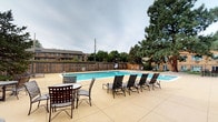 This outdoor amenity space centers around a generously sized rectangular swimming pool with clear, blue water, providing an inviting retreat for recreation and relaxation. The pool is surrounded by a broad, smooth concrete sun deck that offers ample room for lounging, furnished with multiple reclining chairs and a dining-style table and chair set — ideal for socializing or unwinding outdoors. The area is enclosed by a sturdy wooden privacy fence, creating a secure and semi-private atmosphere. Mature pine trees rise within and around the perimeter, providing natural shade and a sense of established, serene landscaping. The surrounding grounds feature low-maintenance gravel and rock landscaping accented by boulders and ornamental stones, complementing the brick exterior of the adjacent building. A gated iron fence entry and concrete walkways provide structured access to the space. The brick building exterior, with its distinctive multi-tone pattern, adds a classic architectural character to the surroundings. Overall, this outdoor pool area combines functional amenities with a comfortable, well-kept environment suited for warm-weather enjoyment.