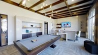 This single-floor entertainment and lounge space is thoughtfully designed to offer a premium social experience. The room's most striking architectural feature is its exposed wood beam ceiling, which adds warmth and character while complementing the modern, neutral palette throughout. A sculptural multi-pendant light fixture cascades from the ceiling, serving as both a functional and artistic focal point above the centerpiece full-size billiards table.

Along one wall, a built-in wet bar features dark cabinetry, open display shelving, a sink, and a full-size refrigerator, creating a fully equipped refreshment station ideal for entertaining. The backsplash incorporates a subtle mosaic tile detail that adds texture to the clean, contemporary aesthetic.

The lounge area is anchored by a large wall-mounted flat-screen TV and comfortable seating, providing a relaxed viewing and social space. Light-toned stone-effect wall cladding frames the TV wall, enhancing the room's upscale ambiance. Plush patterned carpeting underfoot ties the space together, balancing the sleek cabinetry and polished surfaces. Recessed lighting throughout ensures the room is well-illuminated, making this an inviting and versatile space for recreation and gatherings.