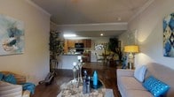 This single-floor residence showcases an open-concept layout that elegantly connects the main living areas, creating a cohesive and functional flow throughout the space. The living room features rich, dark hardwood flooring and is accented with crown molding along the ceiling, lending a refined architectural detail to the interior. Decorative elements, including sculptural centerpieces and an area rug, add character and warmth to the space.

The kitchen is outfitted with warm-toned wooden cabinetry and includes a built-in microwave and range, complemented by a breakfast bar that serves as a natural transition point between the kitchen and the rest of the living area. Overhead fluorescent lighting ensures the kitchen remains bright and functional.

Adjacent to the kitchen, a dedicated dining area accommodates a full dining table and chairs, making it ideal for both everyday meals and entertaining. Lush indoor plants are thoughtfully placed throughout the space, introducing natural greenery that enhances the overall ambiance.

A comfortable seating area anchors the living room, positioned to take in the full expanse of the open floor plan. The combination of hardwood floors, crown molding, and carefully curated décor gives this single-level home a warm, inviting, and well-organized character.