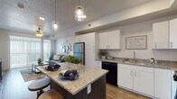 This single-floor residence is designed with a modern, open-concept layout that connects the kitchen, dining, and living areas into one cohesive space. The kitchen is a standout feature, equipped with white shaker-style cabinetry, granite countertops, and a center island with bar seating illuminated by pendant lighting. Black appliances, including a gas range, dishwasher, and stainless steel refrigerator, complement the wood-look flooring that extends throughout the main living areas.

The living room is bright and inviting, benefiting from a sliding glass door that opens to a private balcony offering expansive outdoor views. A ceiling fan adds comfort to the space, which also features a textured ceiling and generous wall space.

Both bedrooms are carpeted and well-proportioned, each accommodating a large bed with room to spare. Natural light filters in through sizable windows fitted with blinds and sheer curtains, offering pleasant exterior views.

The bathroom features white cabinetry, granite countertops, a wide vanity mirror, and a shower-tub combination with a curtain enclosure. Wood-look flooring ties the bathroom's clean, contemporary aesthetic together with the rest of the unit.