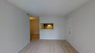 This single-floor residence offers a straightforward and functional layout designed for comfortable everyday living. The main living area is finished with wall-to-wall carpet, creating a warm and inviting atmosphere underfoot. A defining architectural feature of the space is the pass-through window that connects the living area to the kitchen, framed with a light countertop surface and providing a convenient open sightline between the two zones. The kitchen, visible through the pass-through, features dark cabinetry and a classic tile backsplash, lending a traditional character to the cooking space. Adjacent to the living area, a hallway leads further into the residence, where wood-style flooring transitions the aesthetic from the carpeted main space. The entry door is positioned along the right wall of the living area, offering direct and convenient access. Clean white walls and standard ceiling height keep the space feeling bright and neutral, providing a versatile backdrop suited to a variety of furnishing styles. Overall, the layout prioritizes practicality and connectivity between living spaces, making it a functional and approachable single-floor home.