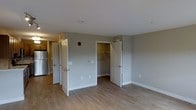 This single-floor residential unit offers a clean, functional layout designed for comfortable everyday living. The main living area features warm hardwood-style flooring and neutral gray walls that create a bright, welcoming atmosphere. White panel doors open to reveal well-organized spaces, including a dedicated closet outfitted with a hanging rod and ample storage capacity, ideal for keeping belongings neatly arranged. The open configuration of the living area allows for flexible furniture placement and a sense of spaciousness. A glimpse into the adjacent kitchen reveals stainless steel appliances, including a refrigerator, complemented by additional cabinetry and functional workspace. The unit is equipped with essential fixtures such as a thermostat panel and smoke detector, reflecting attention to safety and comfort. Crisp white trim and baseboards provide a polished contrast against the soft gray walls, while consistent flooring throughout ties the space together cohesively. The overall design aesthetic is modern and understated, emphasizing practicality and ease of maintenance. Natural light filters through the space, enhancing the clean, airy feel of the interior. This unit is well-suited for those seeking a straightforward, move-in ready living space with thoughtful built-in storage and a neutral palette ready for personalization.
