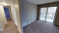 This single-floor residence presents a clean and functional interior characterized by wall-to-wall carpeting in a warm neutral tone, providing a comfortable underfoot feel throughout the living area. The main living space features smooth, gray-painted walls accented by white baseboards, creating a simple yet cohesive aesthetic. A sliding glass door fitted with vertical blinds allows natural light to filter into the room while offering direct access to an outdoor area, enhancing the sense of openness. A floor-level HVAC vent ensures efficient climate control throughout the space. Adjacent to the main room, a hallway leads to additional areas of the unit, with what appears to be a closet featuring louvered doors, adding practical storage options. The textured ceiling is a notable architectural detail consistent throughout the space. Electrical outlets are conveniently positioned along the walls to accommodate various furniture arrangements. Overall, the space maintains a neutral, move-in-ready atmosphere with modest natural light and a straightforward, practical layout suitable for everyday living.