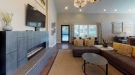 This single-floor community clubhouse offers a thoughtfully designed and generously proportioned gathering space that blends comfort with contemporary style. Soaring vaulted ceilings create an airy, open atmosphere throughout, while rich dark hardwood flooring ties each area together seamlessly.

The main lounge is a standout feature, anchored by a sleek linear fireplace with a mounted flat-screen television above. A large sectional sofa, accented with bold yellow throw pillows, sits atop a woven area rug beneath an elegant chandelier, creating an inviting focal point for relaxation and socializing. Large windows dressed with patterned drapes frame views of the outdoor surroundings, flooding the space with natural light.

A dedicated seating nook near the windows features stylish leather accent chairs and a side table with a decorative lamp — ideal for quiet conversation or casual reading. The open-plan kitchenette is well-appointed with a generous white-topped island with bar seating, rich wood cabinetry, glass-front upper cabinets, a stainless steel sink, and modern pendant lighting. A self-serve refreshment station adds a welcoming, functional touch.

A workstation area with desk seating provides a productive space within the communal layout. Throughout, neutral wall tones, recessed lighting, and carefully selected décor contribute to a polished, modern ambiance suited for both community events and everyday use.